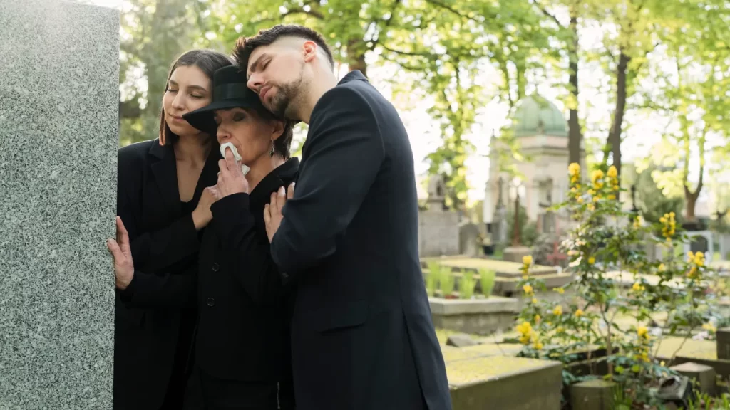 Familiares vestidos de preto se abraçando em frente a um túmulo durante visita ao cemitério.