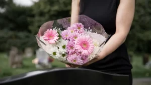 Pessoa segurando um buquê de flores rosas e brancas em frente a um túmulo em cemitério, em gesto de homenagem.