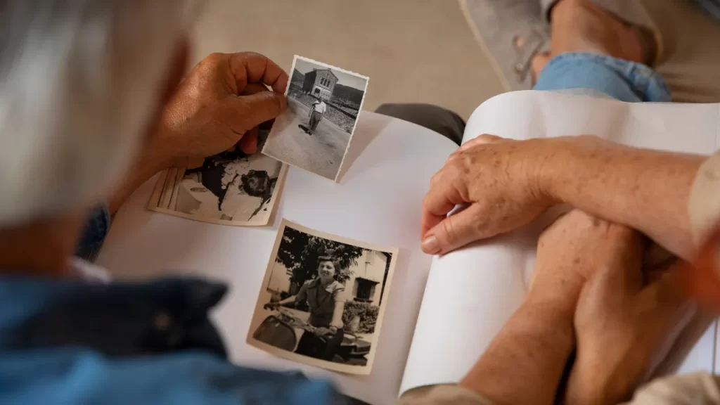 Idoso folheando álbum de fotos antigas em preto e branco, segurando uma fotografia enquanto recorda memórias afetivas.